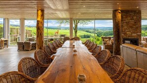 A long wooden table with wicker chairs, a stone fireplace, and a view of a green landscape.