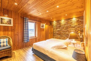A wooden-paneled room with a bed, a wicker chair, a stone wall, and a window with a checkered curtain. - FOREST HILLS LODGE (19 pers) (Stoumont)