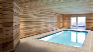 A modern indoor swimming pool with wooden paneling on the walls and ceiling, a concrete floor, and a large window providing a view of the outdoors.