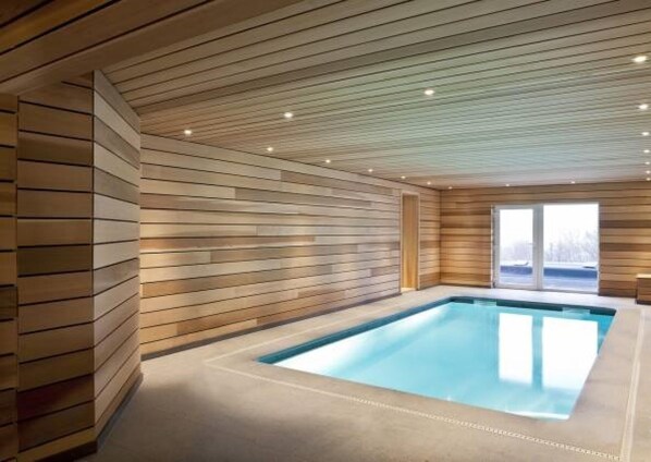 A modern indoor swimming pool with wooden paneling on the walls and ceiling, a concrete floor, and a large window providing a view of the outdoors. - FOREST HILLS LODGE (19 pers) (Stoumont)