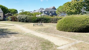 A well-maintained garden with a paved path, a bicycle, and a bench. In the background, there are houses with sloped roofs and a clear blue sky.