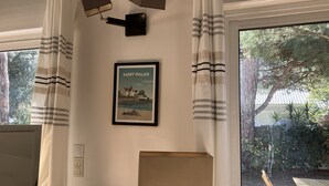 Una habitación con un gran ventanal, cortinas, un cuadro enmarcado de Saint-Palais, una lámpara y una mesa con sillas afuera.