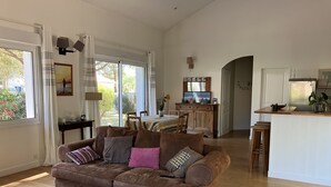 Un salón espacioso con un sofá marrón, una mesa de centro de madera y una zona de comedor con mantel a rayas.