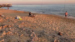 Una playa con gente pescando, un muelle al fondo y un enfriador sobre la arena.