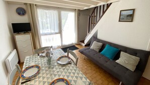 A dining area with a table set for two, a sofa, a TV, and a staircase. - KERNEVEST - Jardin, Tennis, Plage 600m - DT185 (Saint-Philibert)