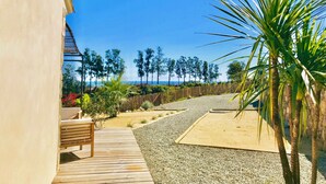 Une terrasse en bois mène à une zone sablonneuse où se trouvent un palmier, un banc et d’où l’on peut admirer l’océan et les arbres.