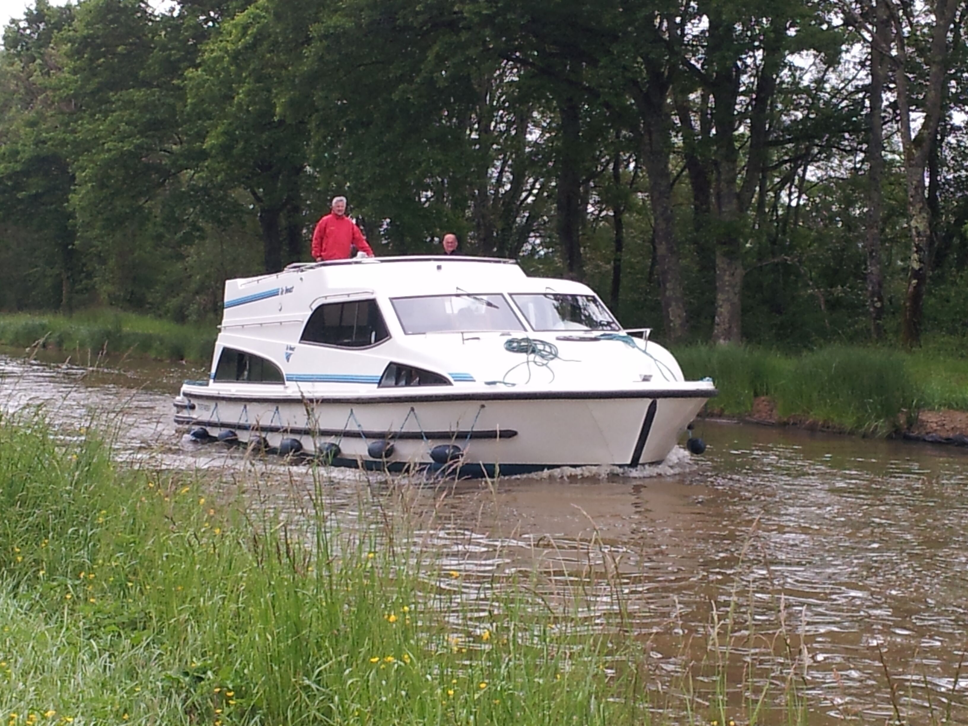 Una lancha blanca con cabina y dos personas a bordo, navegando por un río rodeado de vegetación.