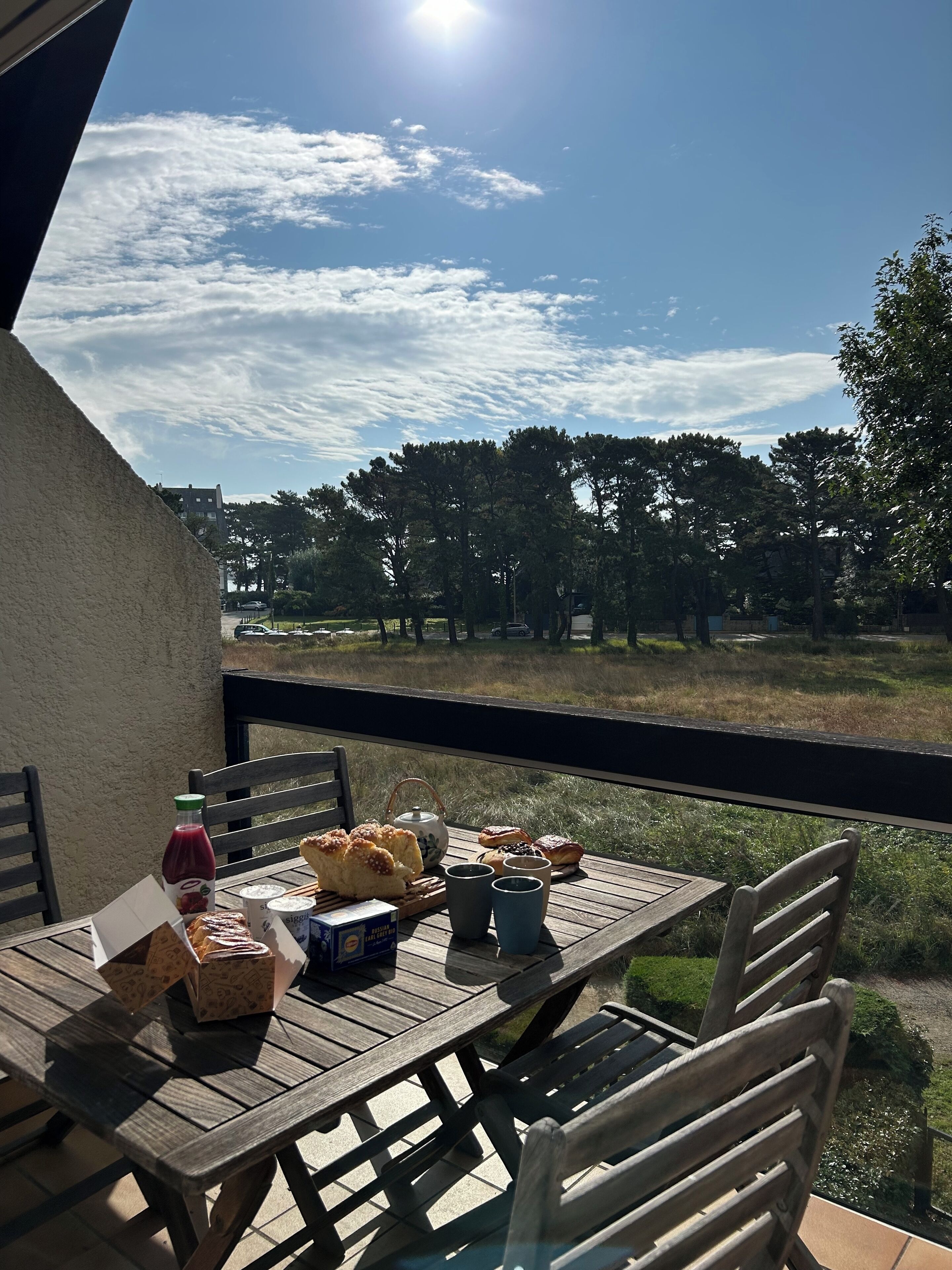 Una mesa de madera con alimentos para el desayuno y una botella de jugo en un balcón con vistas a un área verde con árboles.