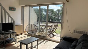 Ein Wohnzimmer mit Fernseher, einer Couch, einem Tisch und einem Sessel. Eine Glastür öffnet sich zu einer Terrasse mit Tisch und Stühlen.