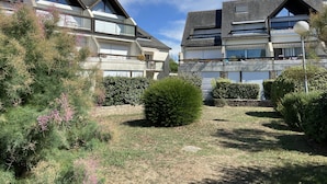 Um edifício residencial com telhado inclinado, várias janelas e um jardim bem cuidado com arbustos e uma pequena árvore.