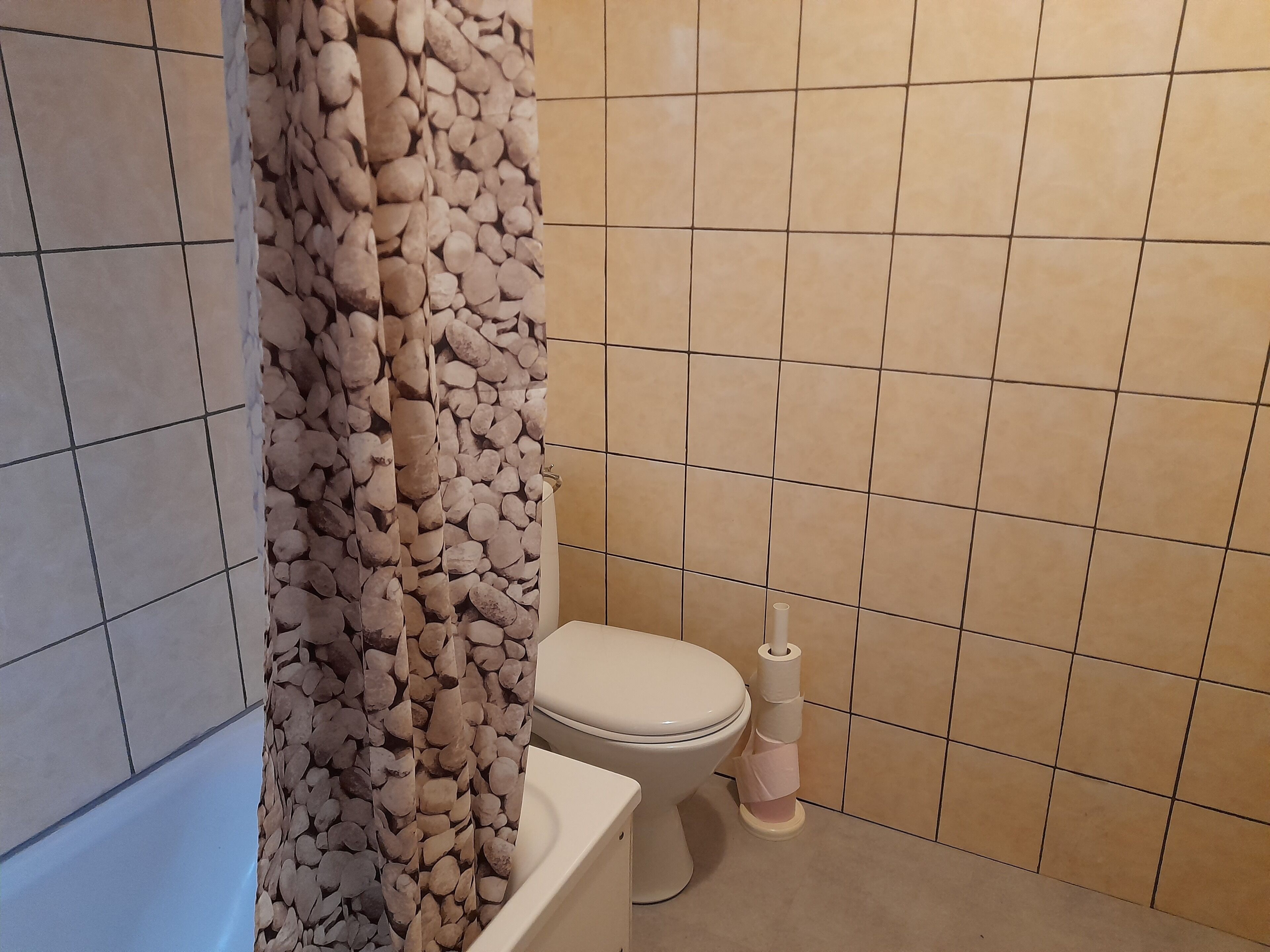 A bathroom with beige tiles, a toilet, and a shower curtain with a pebble pattern.