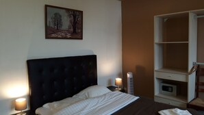Un dormitorio con una cama grande, una mesita de noche con lámpara, un buró con lámpara, un cuadro en la pared y un armario.