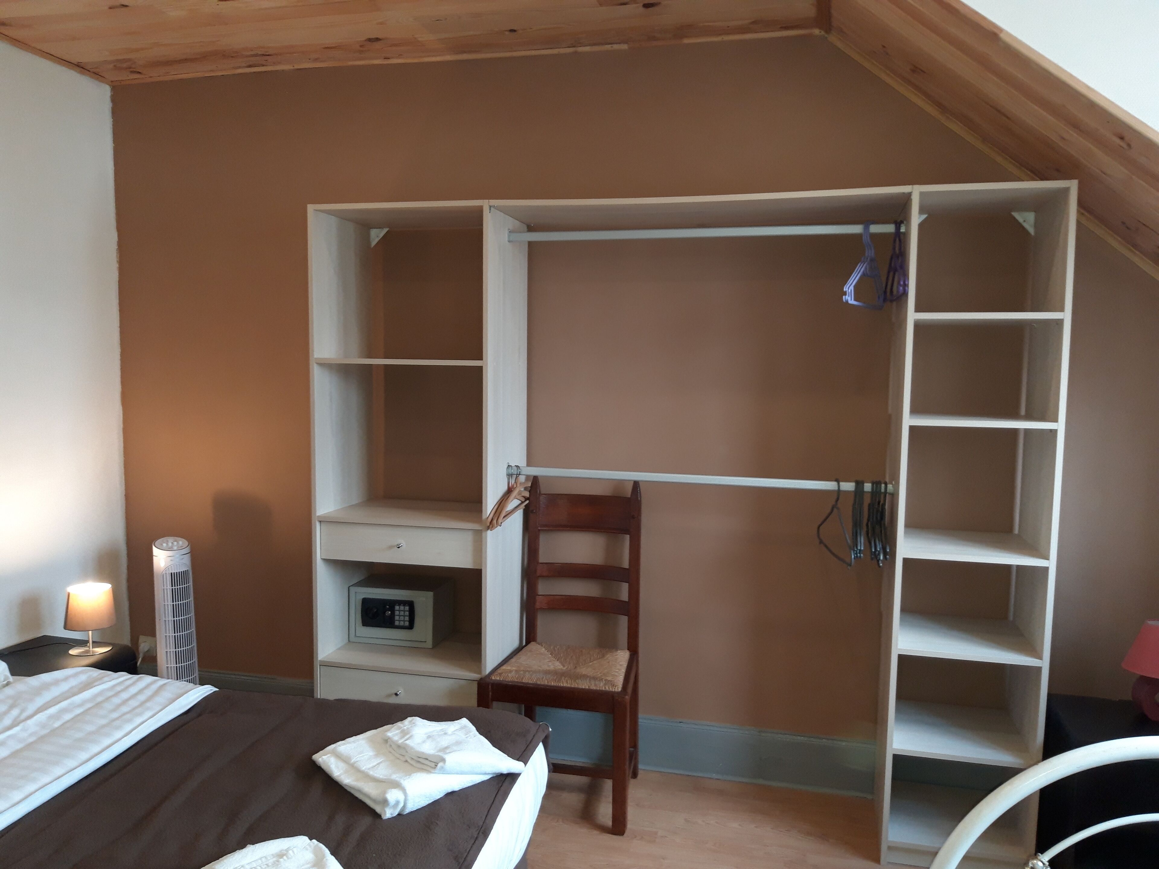Une chambre à coucher comprenant un lit, une chaise en bois, un bureau avec une lampe, une armoire et une petite étagère avec une radio.