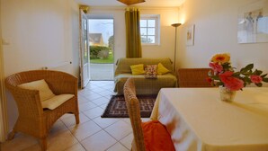Ein Zimmer mit Korbsessel, Sofa mit gelben Kissen, Esstisch mit Blumenvase und Blick ins Freie durch eine Tür.