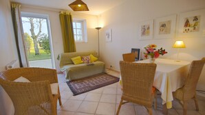 Ein gemütliches Zimmer mit einer Couch, Korbstühlen, einem Esstisch mit Blumenvase und Bildern an der Wand.