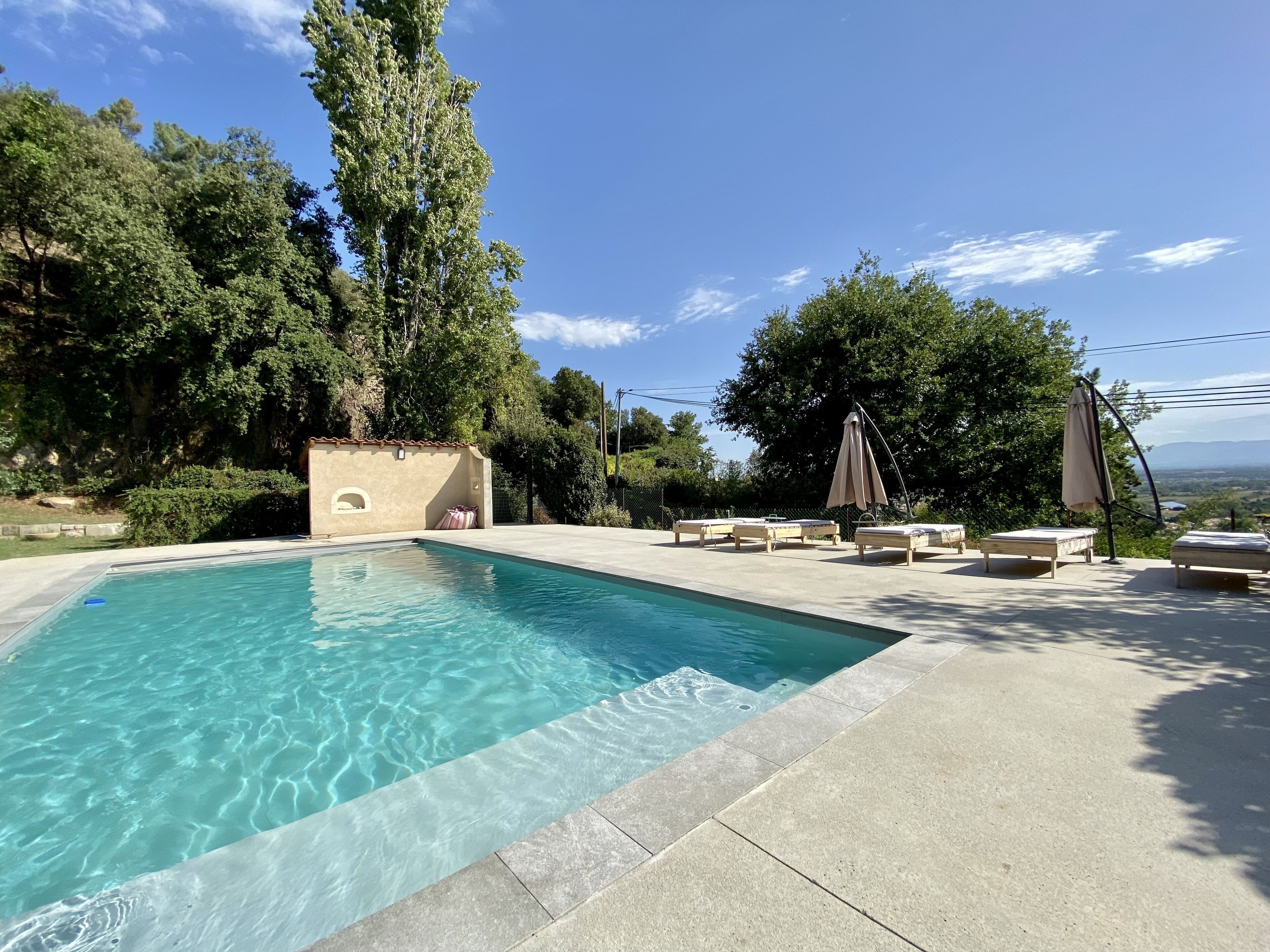A swimming pool with lounge chairs and umbrellas, surrounded by trees and a view of the landscape.