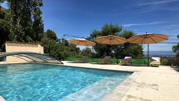 Ein Schwimmbecken mit klarem Wasser, umgeben von Liegestühlen und Sonnenschirmen, mit Bäumen und blauem Himmel im Hintergrund.