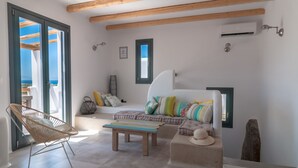 Ein modernes Wohnzimmer mit einer Couch, einem Holzstuhl und einem kleinen Tisch. Es gibt Fenster mit Holzbalken darüber und eine Klimaanlage an der Wand.