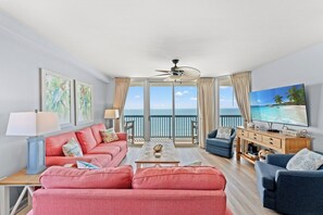 A living room with a sofa, armchairs, a coffee table, a TV, and a balcony with a view of the ocean.