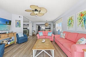 A living room with a TV, a ceiling fan, a pink sofa, a blue armchair, and a wooden coffee table.