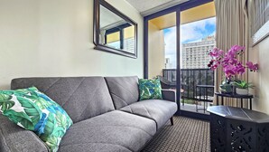 Ein graues Sofa mit gemusterten Kissen, ein kleiner Tisch mit Blumentöpfen und ein Balkon mit Blick auf Gebäude.