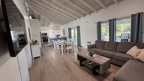 A spacious living area with a wooden floor, a sofa, a coffee table, and a kitchen in the background. - Gated Villa w Private Pool, Outdoor Shower, BBQ • Easy Walk to GraceBay Beach (Providenciales)
