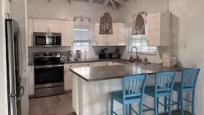 A modern kitchen with white cabinets, stainless steel appliances, a large island with blue chairs, and a ceiling fan. - Gated Villa w Private Pool, Outdoor Shower, BBQ • Easy Walk to GraceBay Beach (Providenciales)