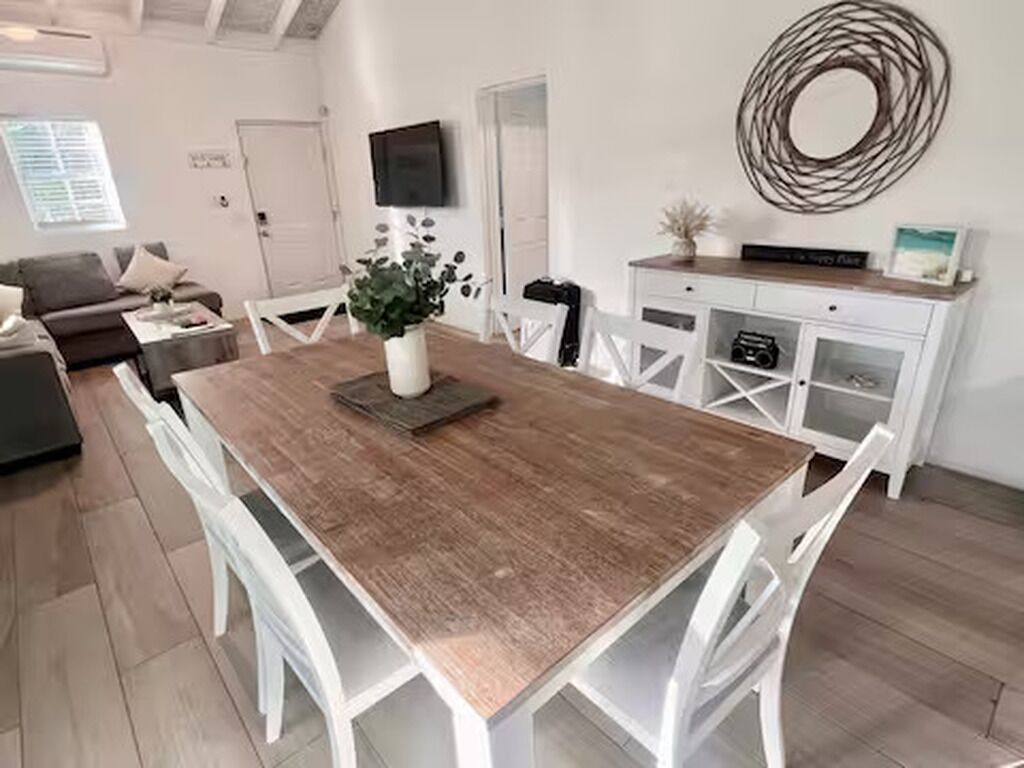 Un coin repas avec une table en bois et des chaises blanches, une plante en pot et un meuble blanc sur lequel est posée une télévision.