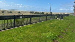 A grassy area with a metal fence, a view of the beach, and a building with balconies. - On the Promenade 1st floor. Spectacular Panoramic Ocean Views, Patio with grassy (Seaside)