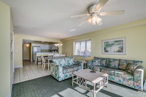 A living room with a sofa, a coffee table, and a ceiling fan. There is a kitchen in the background.
