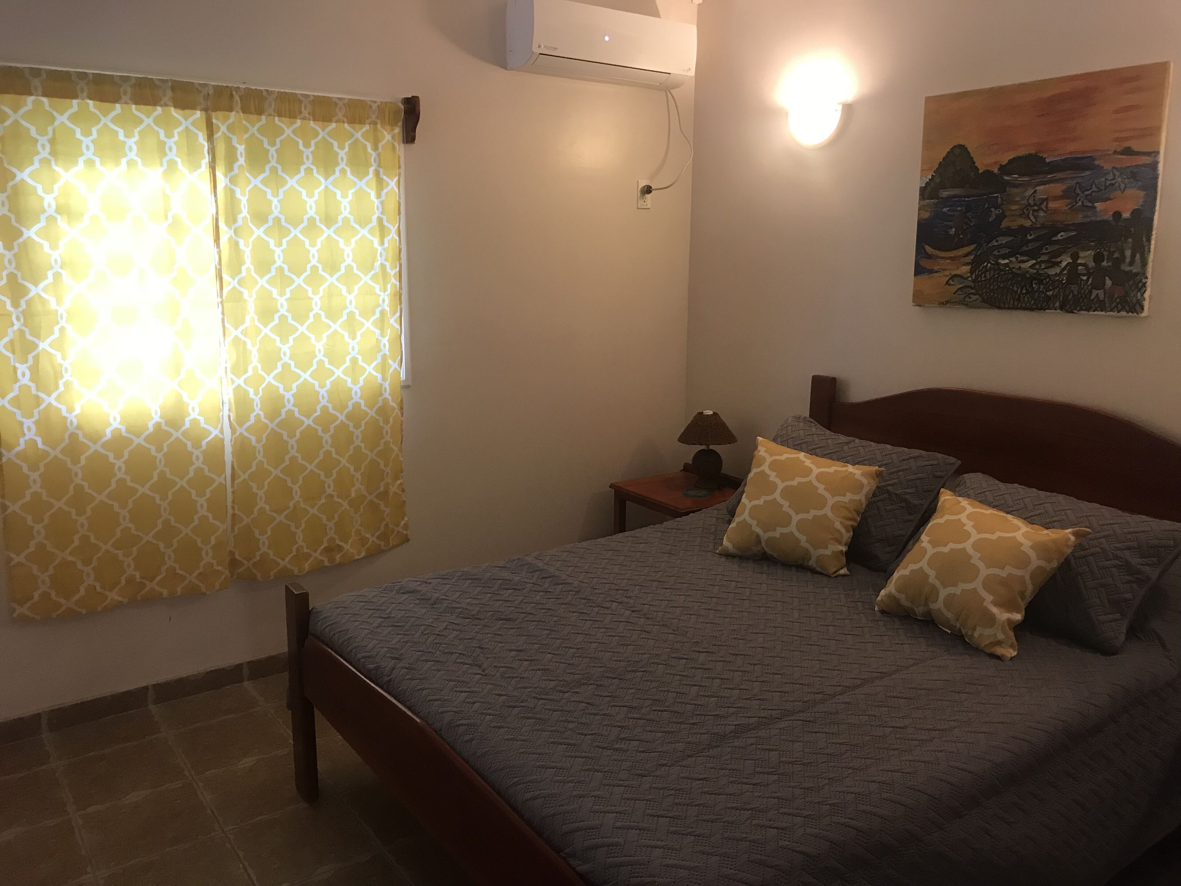 Un dormitorio con cama, mesita de noche, lámpara, aire acondicionado instalado en la pared y un cuadro.