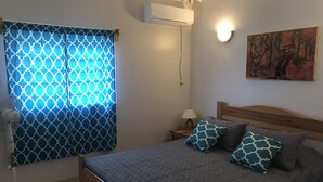 Une chambre à coucher avec un lit, des rideaux à motifs, un climatiseur fixé au mur, une lampe de chevet et un tableau.