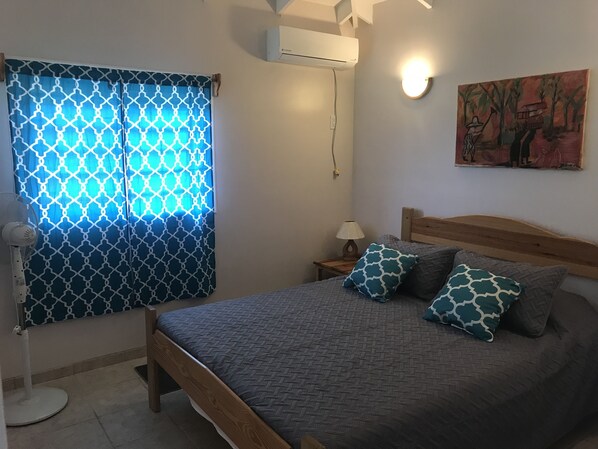 Un dormitorio con cama, cortinas estampadas, aire acondicionado de pared, lámpara de noche y un cuadro.