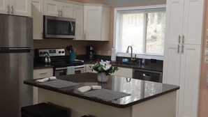 A modern kitchen with white cabinets, a stainless steel refrigerator, a microwave, and a coffee maker. There is a window with blinds, a sink, and two bar stools. - Mountainside Spa With View’s  (Bushkill)