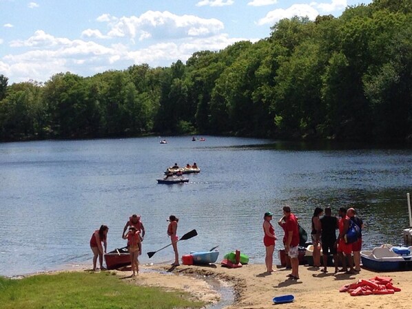Escena junto al lago con gente cerca del agua, botes y un área boscosa al fondo.