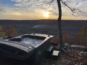 A hot tub in a natural setting with a view of a forested landscape and a sunset in the background.