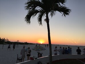 A beach scene with people, a palm tree, and a sunset. - <br>Partial Ocean View 2BR 2BATH<br>Sleeps 4. (Fort Myers Beach)