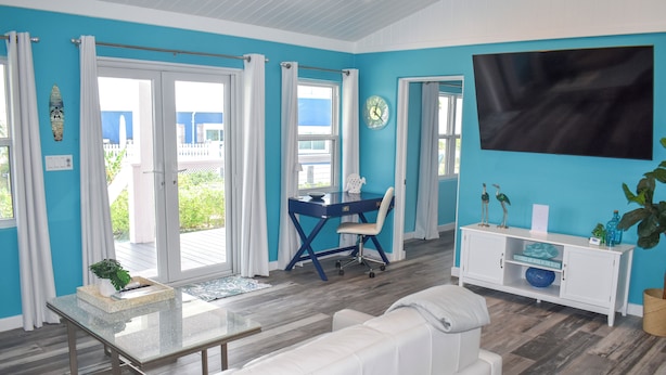 Une pièce lumineuse avec un mur bleu, des meubles blancs, une table basse en verre, un bureau avec une chaise et un téléviseur fixé au mur.