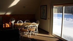 Een eethoek met een houten tafel en stoelen, een ventilator en uitzicht op een besneeerd landschap door een glazen deur.