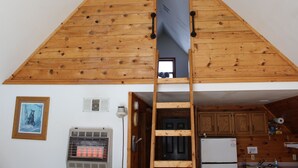 Een houten slaapkamer op zolder met een ladder naar een deur, een klein raam en een kitchenette met kastjes en een koelkast.