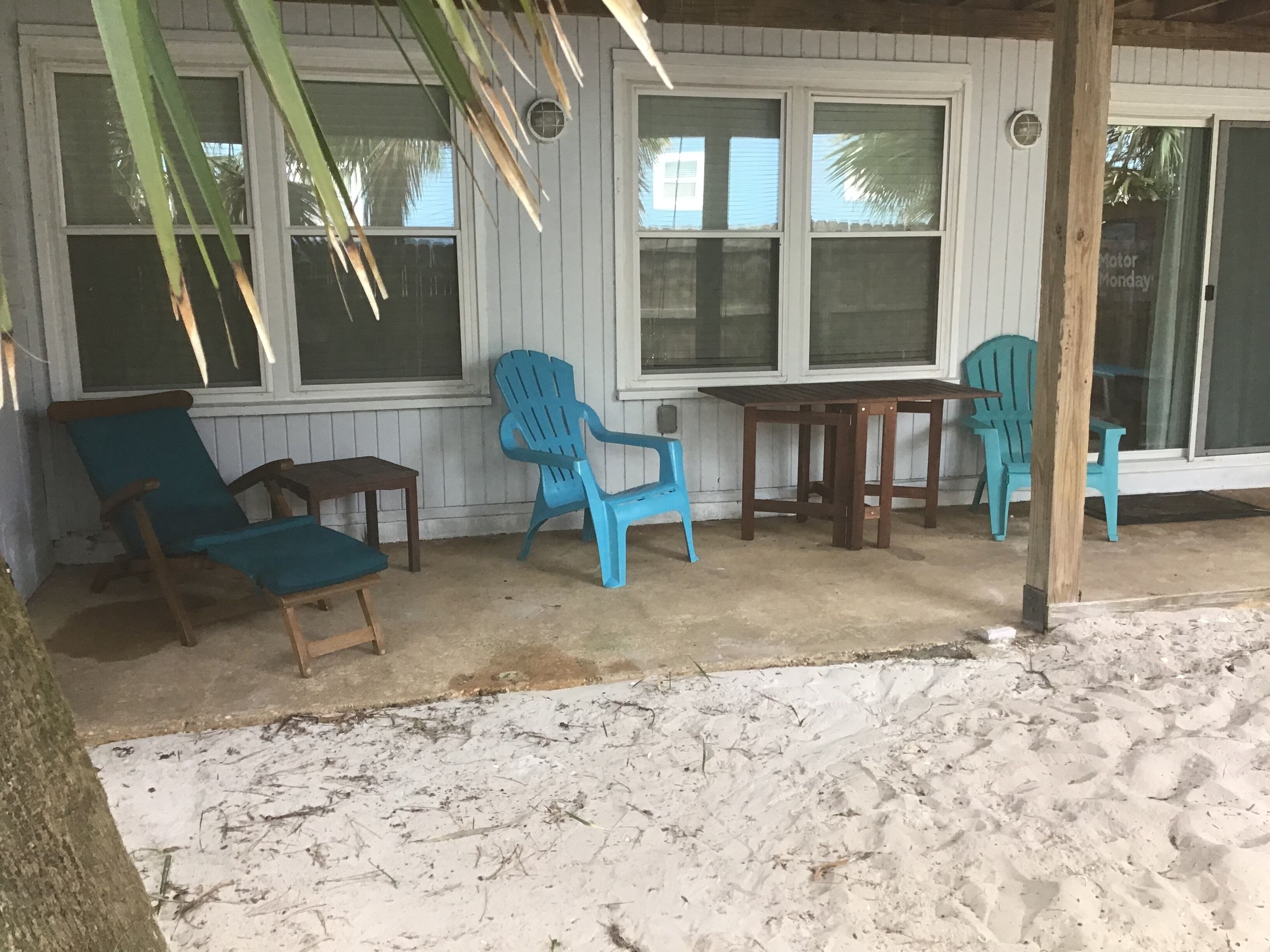 Un espacio al aire libre cubierto con una mesa de madera y cuatro sillas, dos de madera y dos de plástico, cerca de un edificio blanco con ventanas.