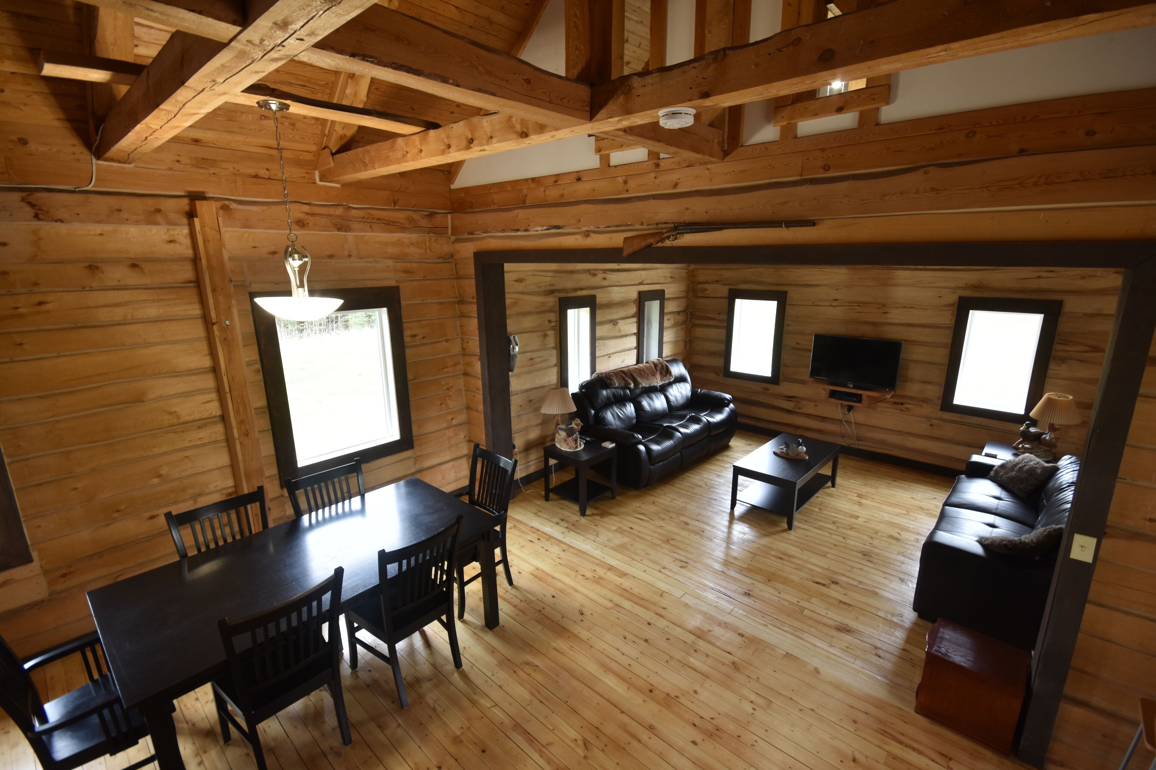 A log cabin interior with a dining area, a black leather sofa, and a television.