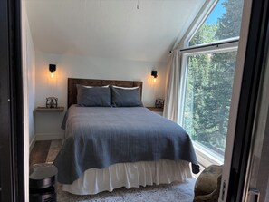 A bedroom with a large window, a bed with a grey blanket, two bedside tables with lamps, and a wooden headboard.