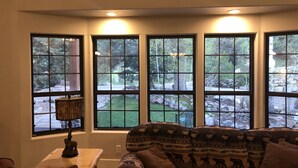 A living room with a sofa, a table with a lamp, and large windows offering a view of a garden.