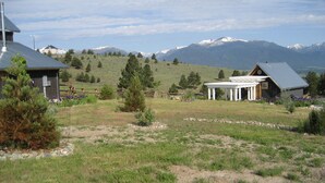 Un paysage rural avec une maison dotée d’un porche et d’un toit en métal, entourée d’une zone délimitée par une clôture, où l’on trouve de l’herbe et des arbres.