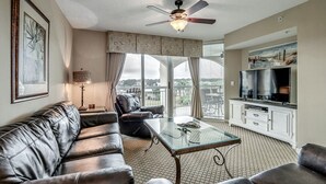 A living room with a leather sofa, a glass coffee table, a ceiling fan, and a kitchen area with a TV. - 3rd Floor Marina View. Hot Tubs, Pool, Near Golf | Barefoot North Tower - 301 (North Myrtle Beach)