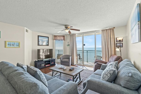 A living room with a TV, sofa, and a balcony with a view of the sea.