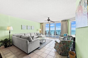 A living room with a grey sofa, a coffee table, and a patterned armchair. There is a ceiling fan, a balcony with a view of the ocean, and a floor lamp.