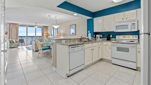 A modern kitchen with white cabinets, a built-in microwave, and a stove. There is a dining area with a table and chairs, and a view of the ocean through large windows. - Upscale Oceanfront Condo, Jacuzzi Tub, Lazy River | North Shore Villas - 405 (North Myrtle Beach)
