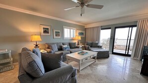 Ein großzügiges Wohnzimmer mit Deckenventilator, grauen Polster-Sofas, einem weißen Couchtisch und Blick durch eine Schiebetür auf einen Außenbereich.